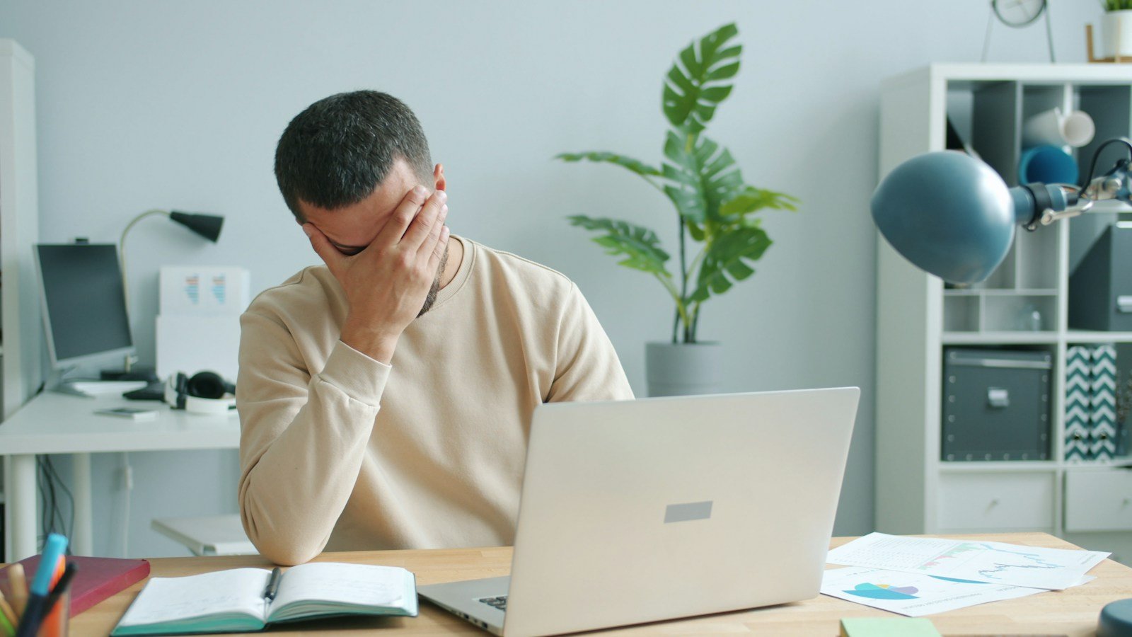 A man sitting at a desk with his hand covering his face, beside a laptop, open notebook, and papers.