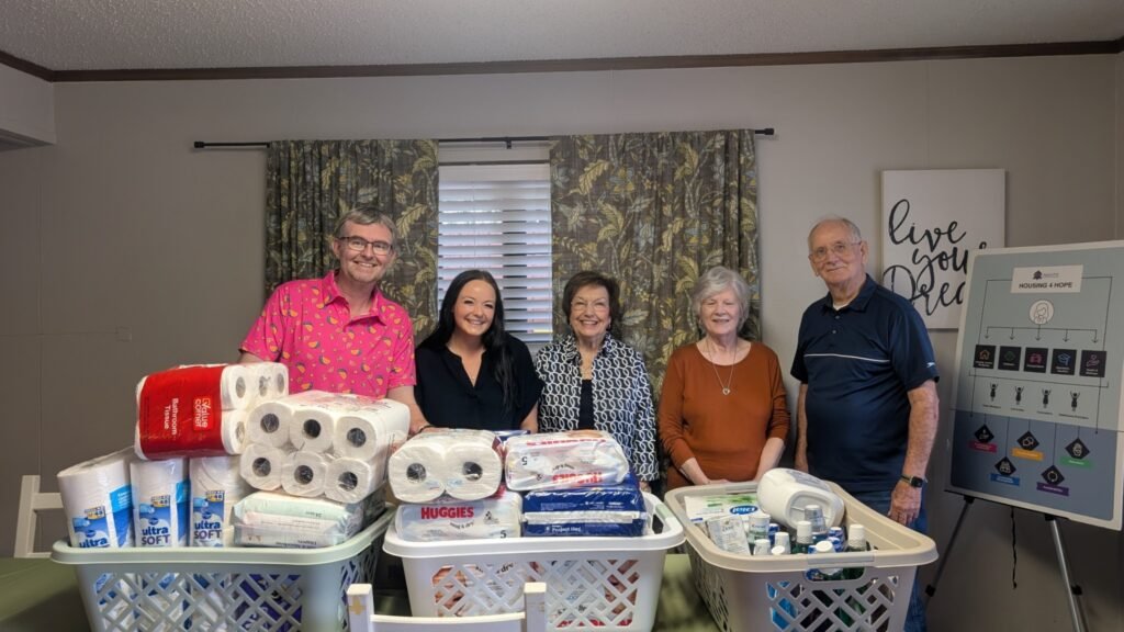 A group with donated household supplies in front of Agape Housing 4 Hope signage.