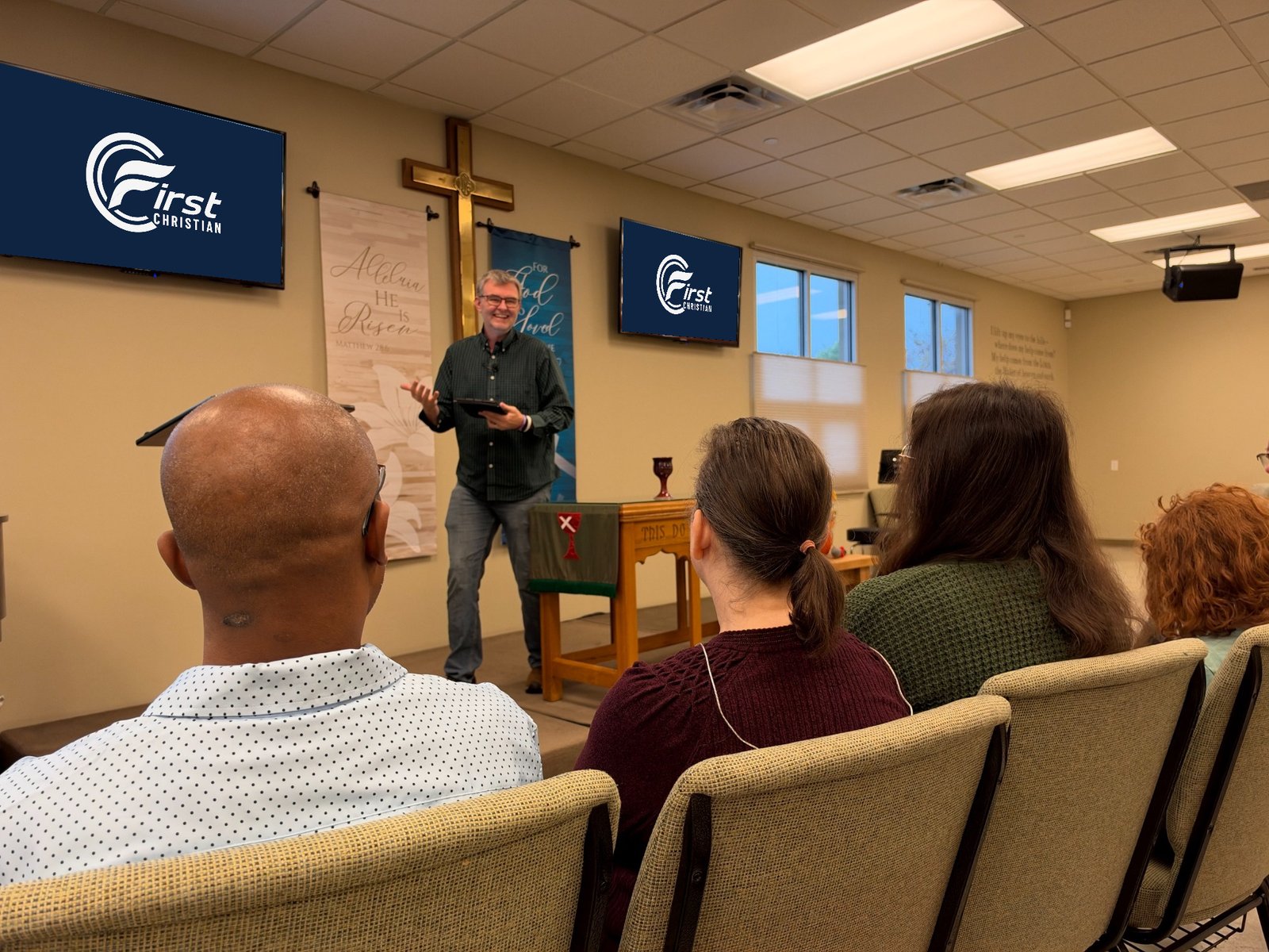 Pastor Chris Fluitt speaking to seated listeners at First Christian Church of Allen, with church logos displayed behind him.